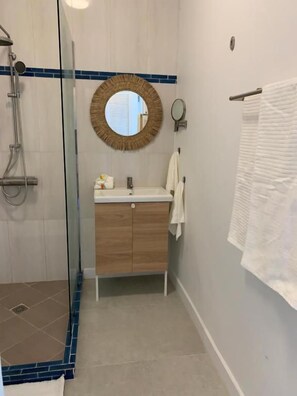 A bathroom with a glass shower enclosure, a wooden vanity, a round mirror, and a towel rack. - Naturalis Oceanview Residences (Victoria)