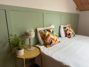 A bed with white bedding, two pillows with a chevron pattern, a bedside table with a lamp and a potted plant, and a green headboard.