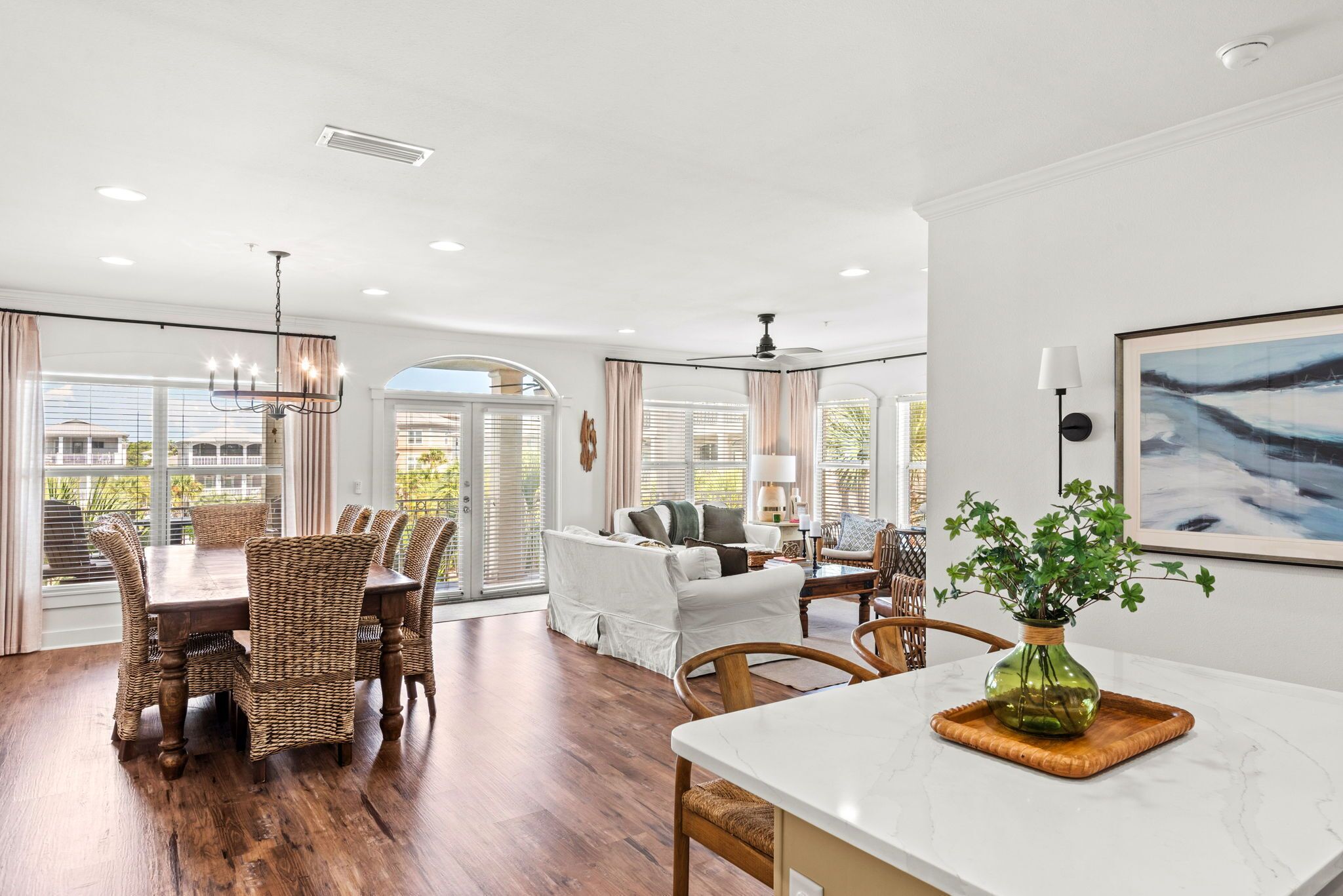 A spacious living area with a dining table, wicker chairs, a sofa, and a large window with blinds.