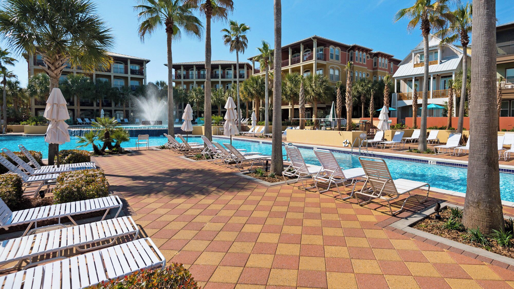A resort pool area with multiple lounge chairs and umbrellas, surrounded by palm trees and a building with balconies.