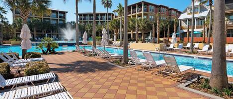 A resort pool area with multiple lounge chairs and umbrellas, surrounded by palm trees and a building with balconies.