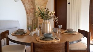 A round dining table with a rustic wooden finish, surrounded by cushioned chairs. A vase with flowers is placed on the table, and there are plates and glasses set for a meal.