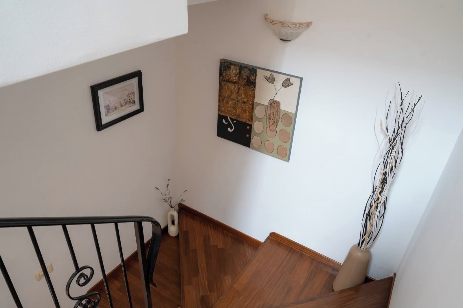 A staircase with a wooden handrail and a black metal railing, leading to a landing with a framed picture and a vase with branches.