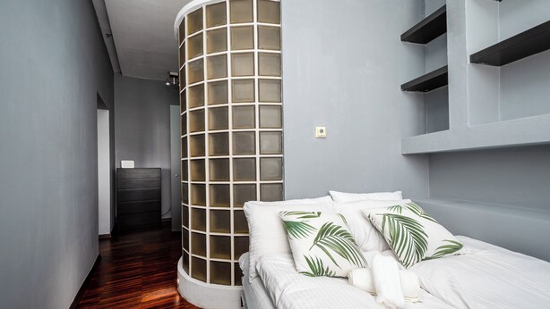 Una habitación de hotel moderna con un separador de vidrio curvo, una cama con ropa blanca y almohadas con forma de hoja de palmera, y estanterías integradas.