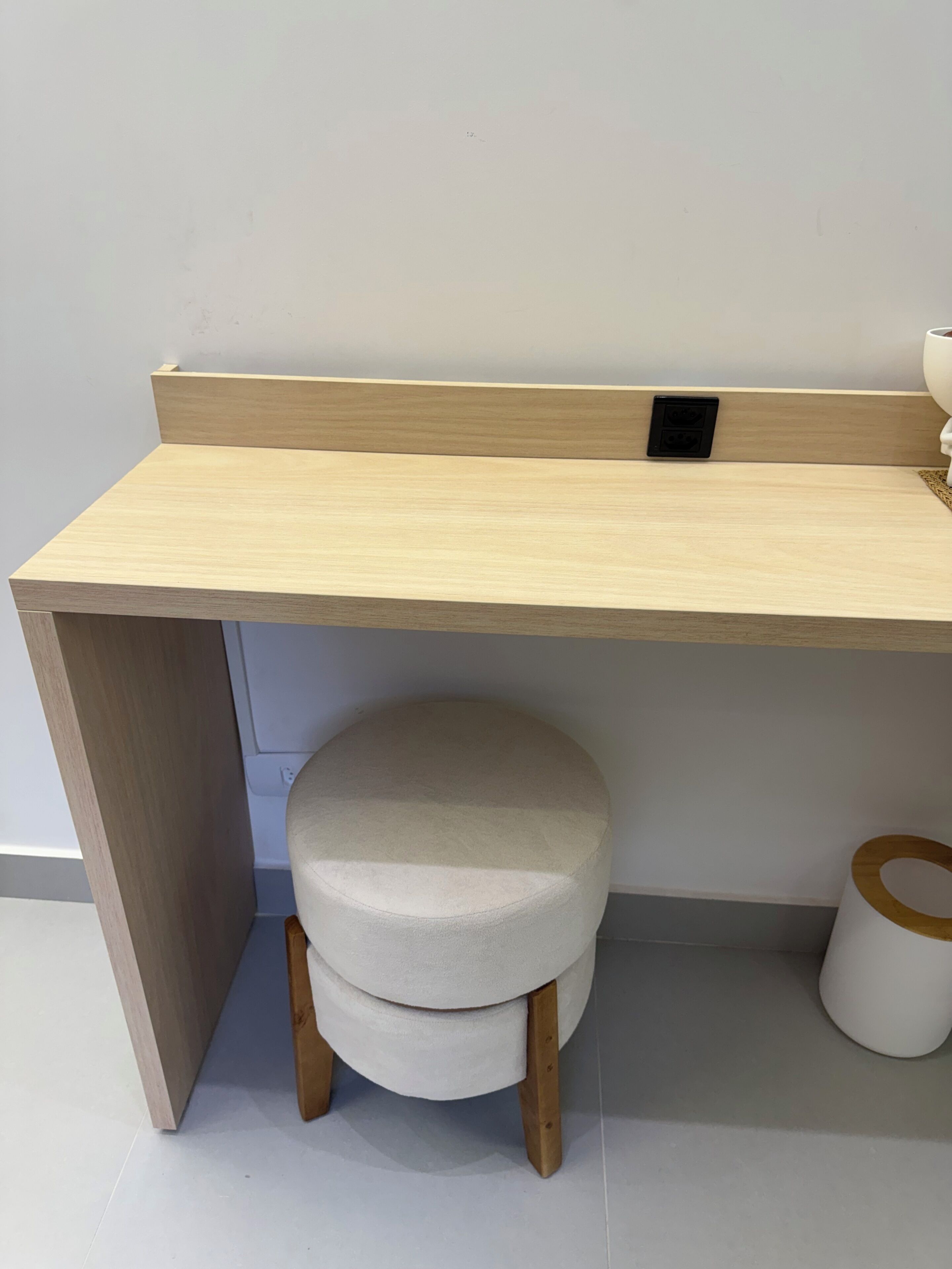 Una scrivania in legno con uno sgabello rotondo sottostante, una presa di corrente e un cestino bianco a lato.