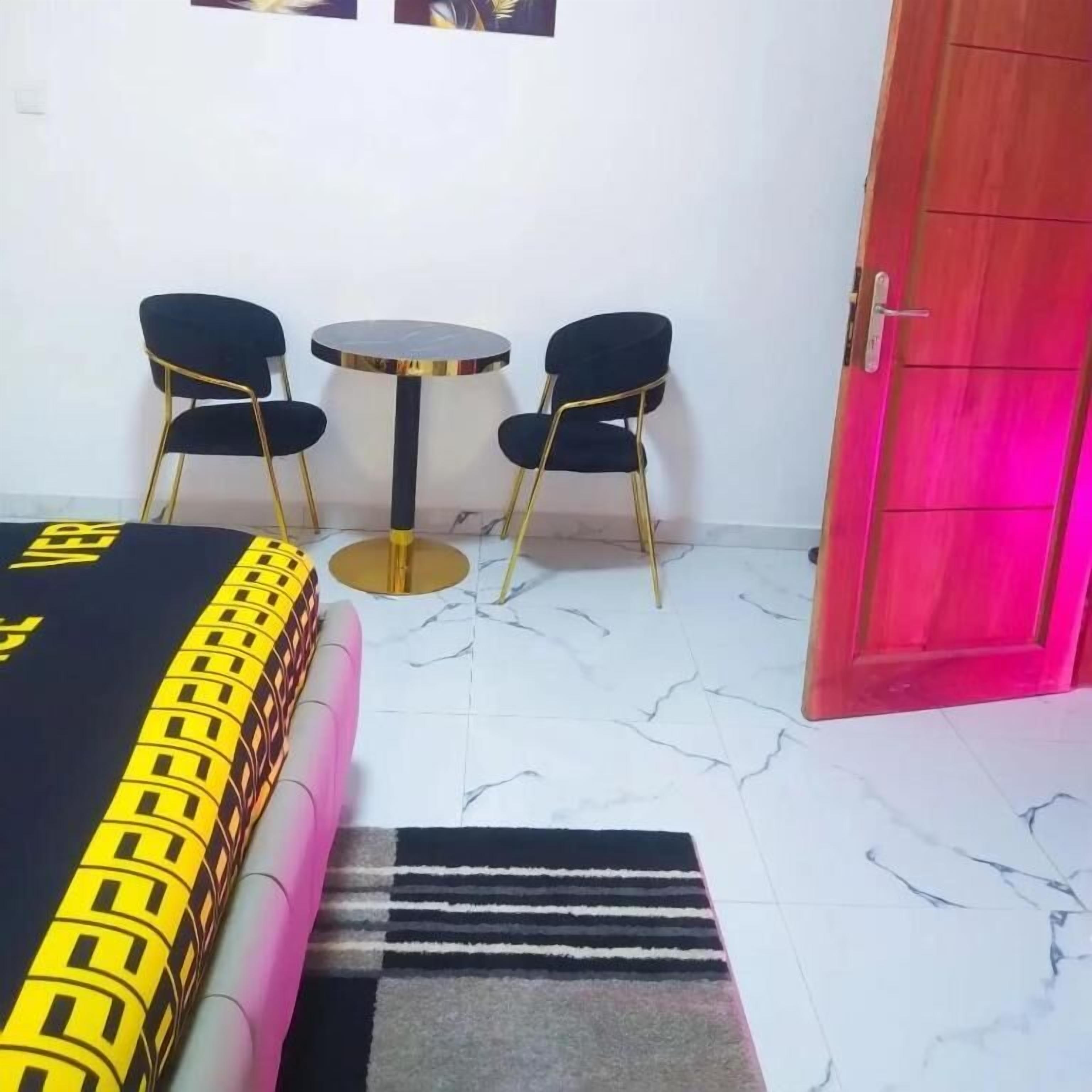 A hotel room with a bed, two chairs, a small table, a red door, and a marble floor.