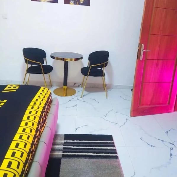 A hotel room with a bed, two chairs, a small table, a red door, and a marble floor.