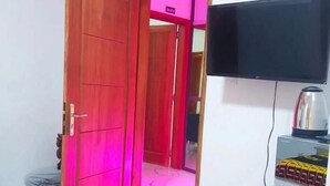 A room with a wooden door, a television on a stand, and a red-lit hallway. - Chambre SCAT urbam (Dakar)
