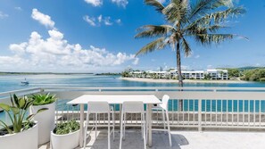 A balcony with a table and chairs overlooking a pool and a beach with a resort in the background. - Bella Vista Waterfront Luxury (Noosaville)