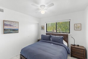 Un dormitorio con una cama, una mesita de noche, un ventilador de techo y una ventana con vistas a árboles.