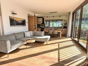 Ein modernes Wohnzimmer mit einem Ecksofa, einem kleinen Couchtisch und einem Essbereich mit Tisch und Stühlen. Im Hintergrund ist eine Küche mit einem großen Fenster zu sehen.