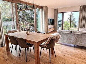 Ein moderner Essbereich mit Holztisch und -stühlen, ein Balkon mit Blick auf Bäume und ein Wohnbereich mit Sofa und Fernseher.