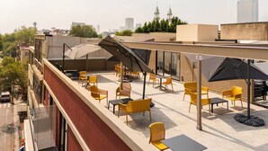 Eine Dachterrasse mit gelben Stühlen und Tischen, einem überdachten Bereich und Blick auf die Stadtlandschaft.