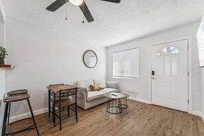 A room with a white door, a ceiling fan, a round mirror, a sofa, a coffee table, and a dining table with chairs. - Downtown Chandler Escape – Walkable Location (Chandler)
