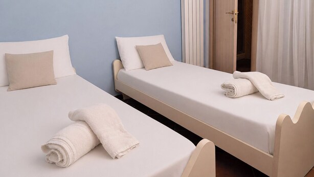 Dos camas individuales con sábanas blancas y almohadas color beige en una habitación con paredes azul claro.