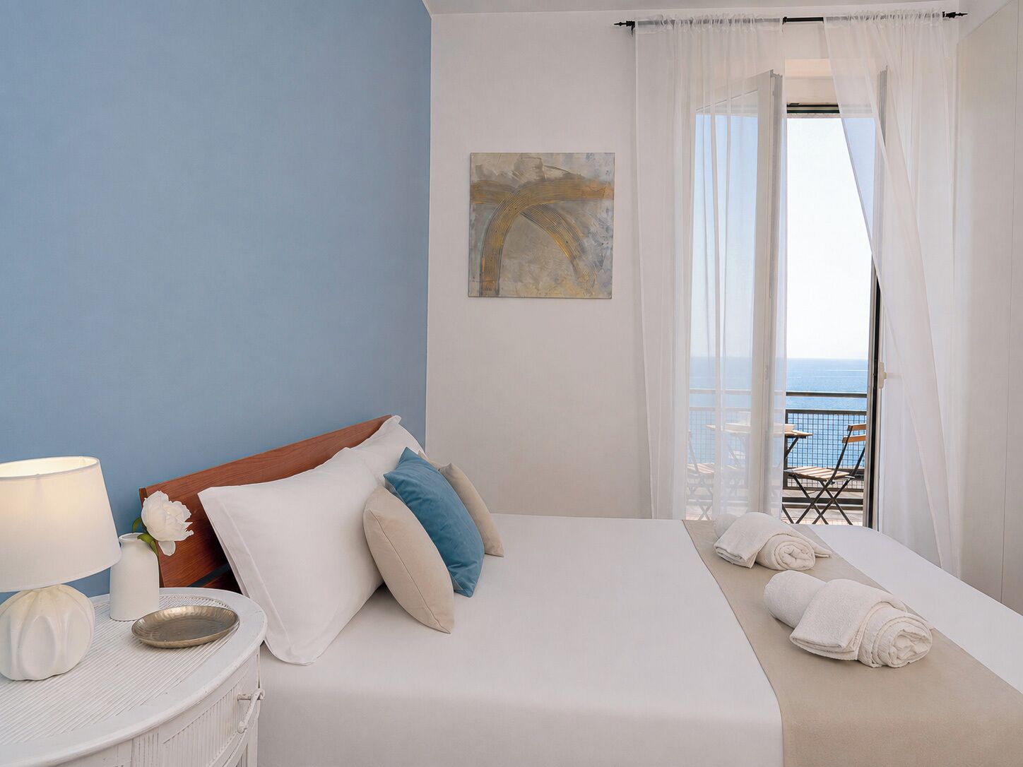 A hotel room with a bed, a bedside table with a lamp, a painting on the wall, and a view of the sea through the window.