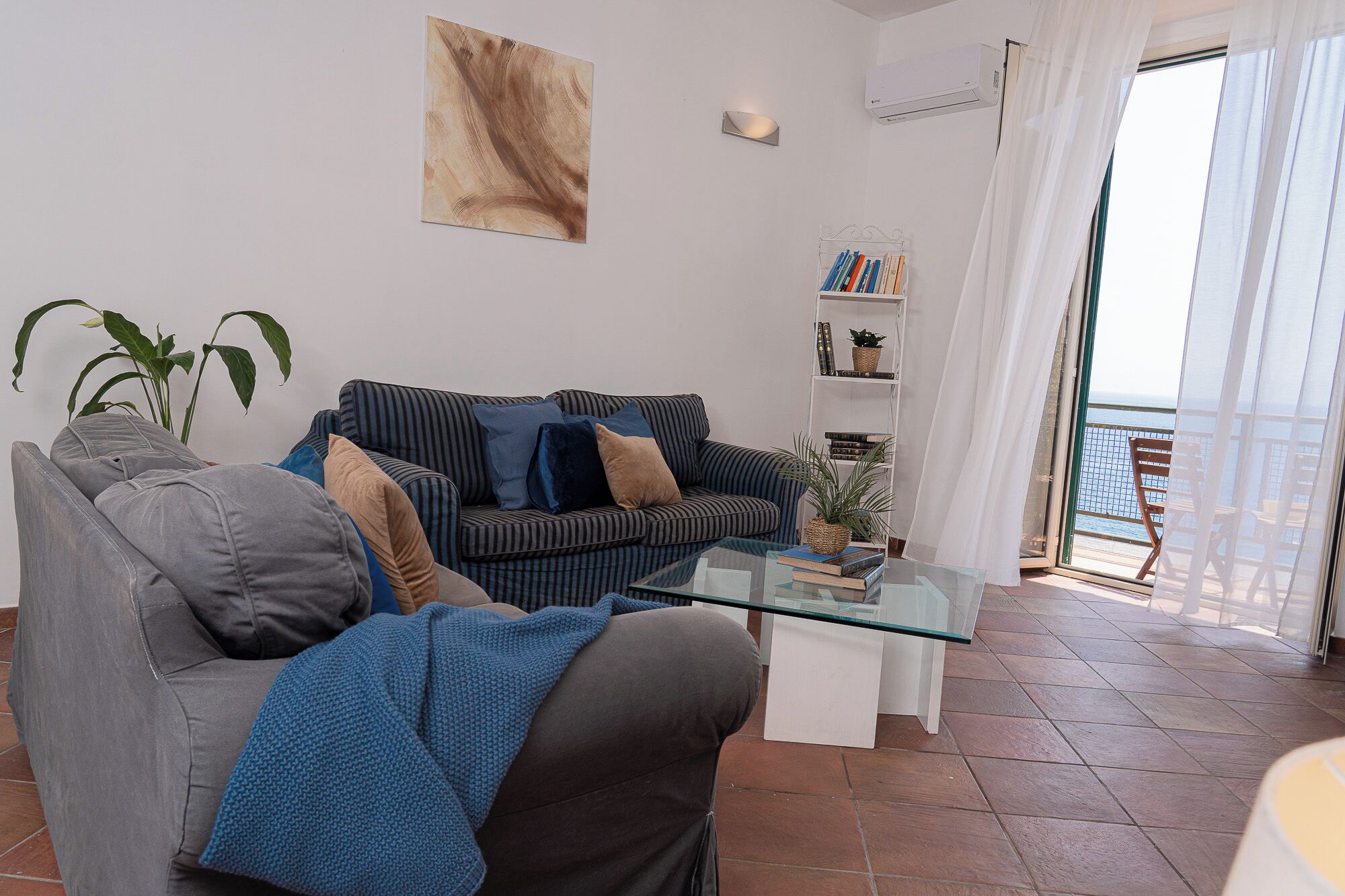 Un salon comprenant un canapé, un fauteuil et une table basse en verre. On y trouve également une bibliothèque avec des livres et une plante, ainsi qu’un tableau accroché au mur.