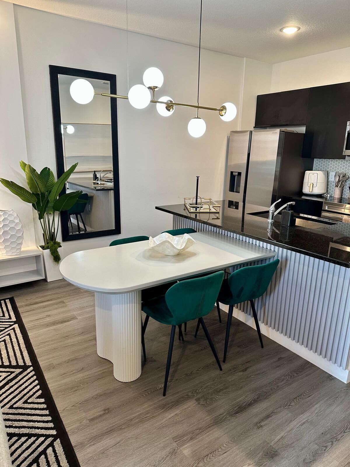 Une cuisine moderne dotée d’un îlot central, d’un coin repas avec une table blanche et des chaises turquoise, et d’un grand miroir surmonté d’un luminaire.