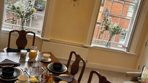 Une salle à manger avec une table dressée pour quatre personnes, un lustre et une vue sur un bâtiment en briques, visible à travers de grandes fenêtres.