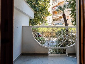 Ein Balkon mit weißem Metallgeländer, Fliesenboden und Blick auf eine Wohngegend mit Bäumen und parkenden Autos.