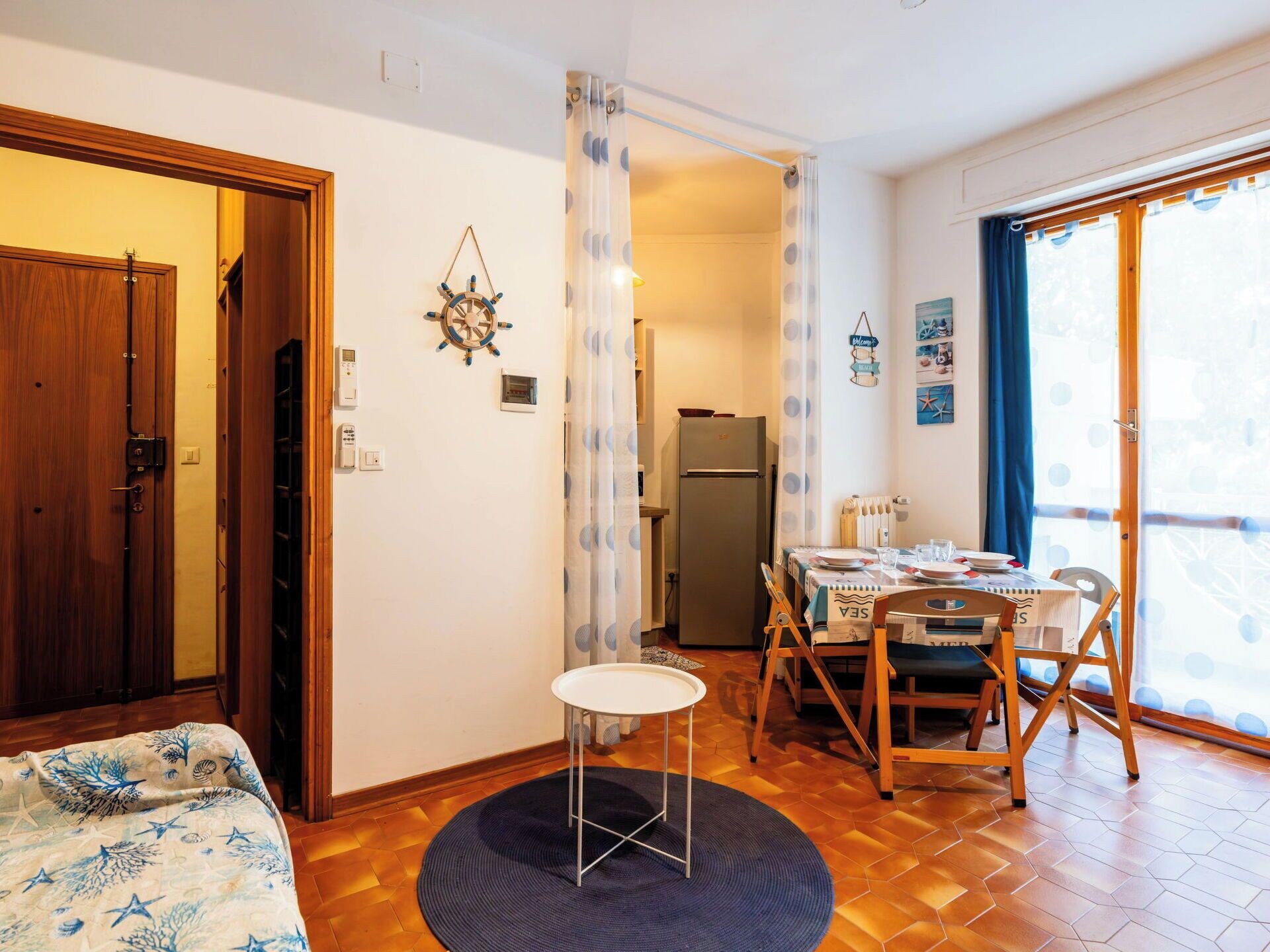 A compact room with a bed, a dining table set for two, a refrigerator, and a view through a door.