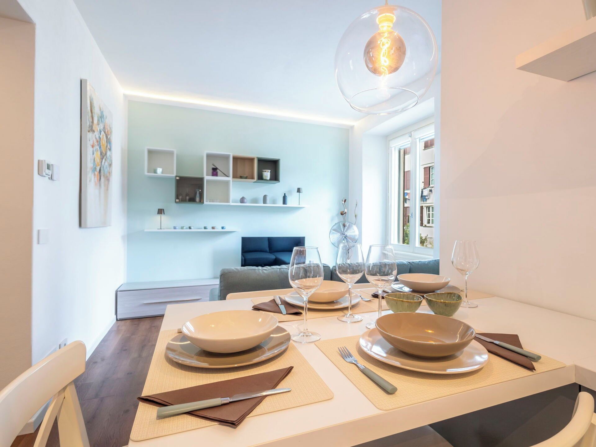 A dining area with a table set for two, a sofa in the background, and a modern light fixture.