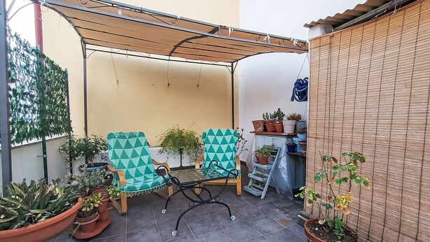 Ein überdachter Patio-Bereich mit zwei Stühlen, Topfpflanzen und einem Holztisch.