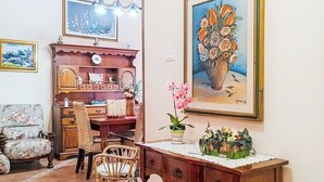 A room with a wooden desk, wicker chairs, a bookshelf, and a large framed painting of flowers.
