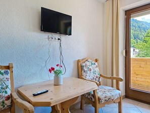 Ein Zimmer mit Holztisch, zwei Stühlen, einem an der Wand befestigten Fernseher und einem Fenster mit Blick ins Grüne.