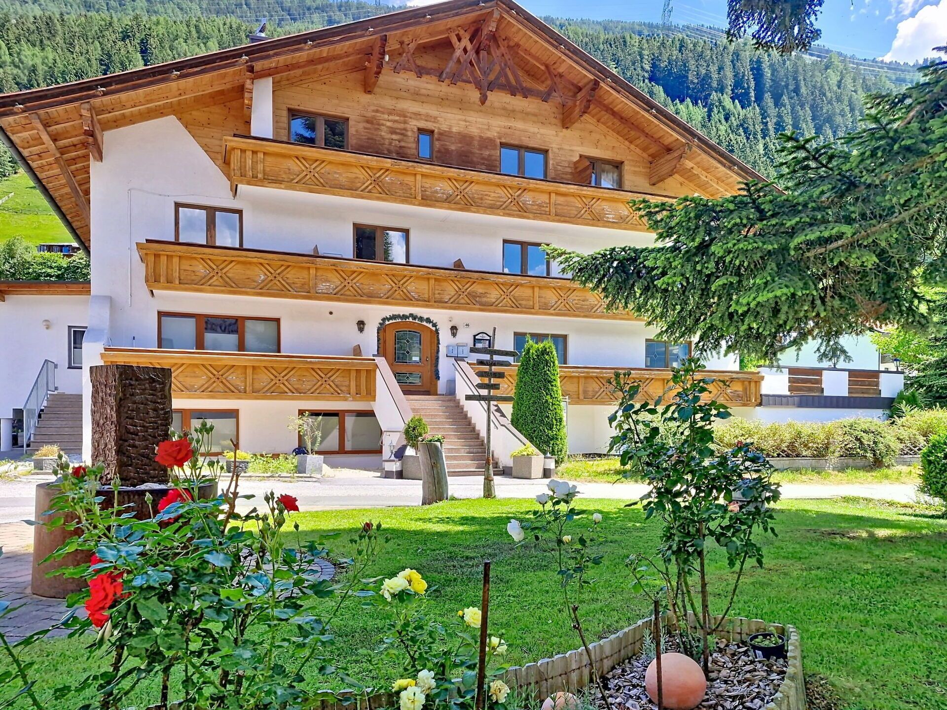 A two-story wooden building with a staircase leading to the entrance, surrounded by a well-maintained garden with flowers and a clear view of the mountains in the background.