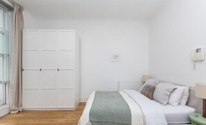 A bedroom with a bed, bedside table, lamp, wardrobe, and a window with curtains. - Regents Park views - light filled studio, 1 min to Baker St tube. (London)