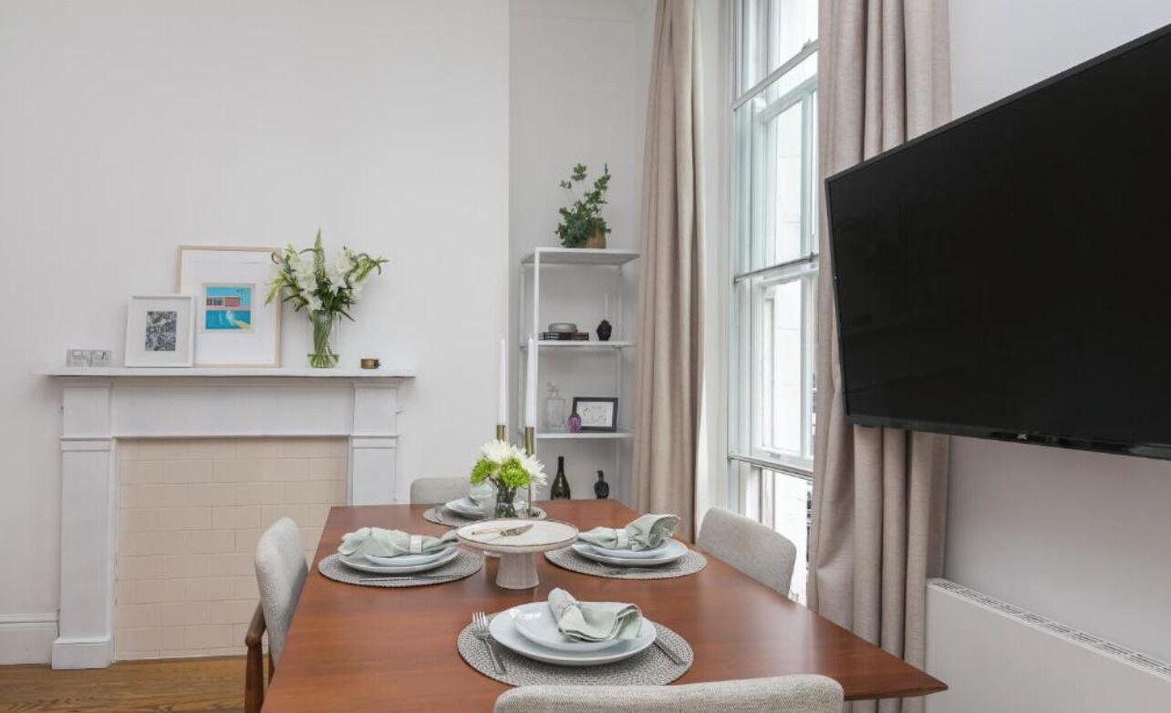 Een eetgedeelte met een houten tafel voor vier personen, een aan de muur bevestigde televisie, en een witte schoorsteenmantel met ingekaderde foto's en een bloevas.