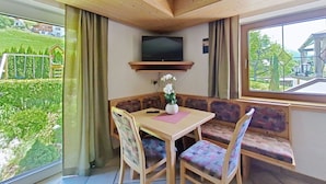 Ein Zimmer mit Holzboden, einem an der Wand befestigten Fernseher, einem Tisch mit Stühlen, einer Bank und einem Fenster mit Blick auf eine grüne Landschaft.