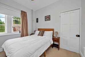 A bedroom with a bed, a nightstand, a lamp, a door, and a window with curtains. - The Jewel In Bury (Bury St Edmunds)