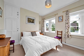 Ein Schlafzimmer mit einem Bett, einem Holznachttisch, einem Sessel, einem Schreibtisch und einem Fenster mit Vorhängen.