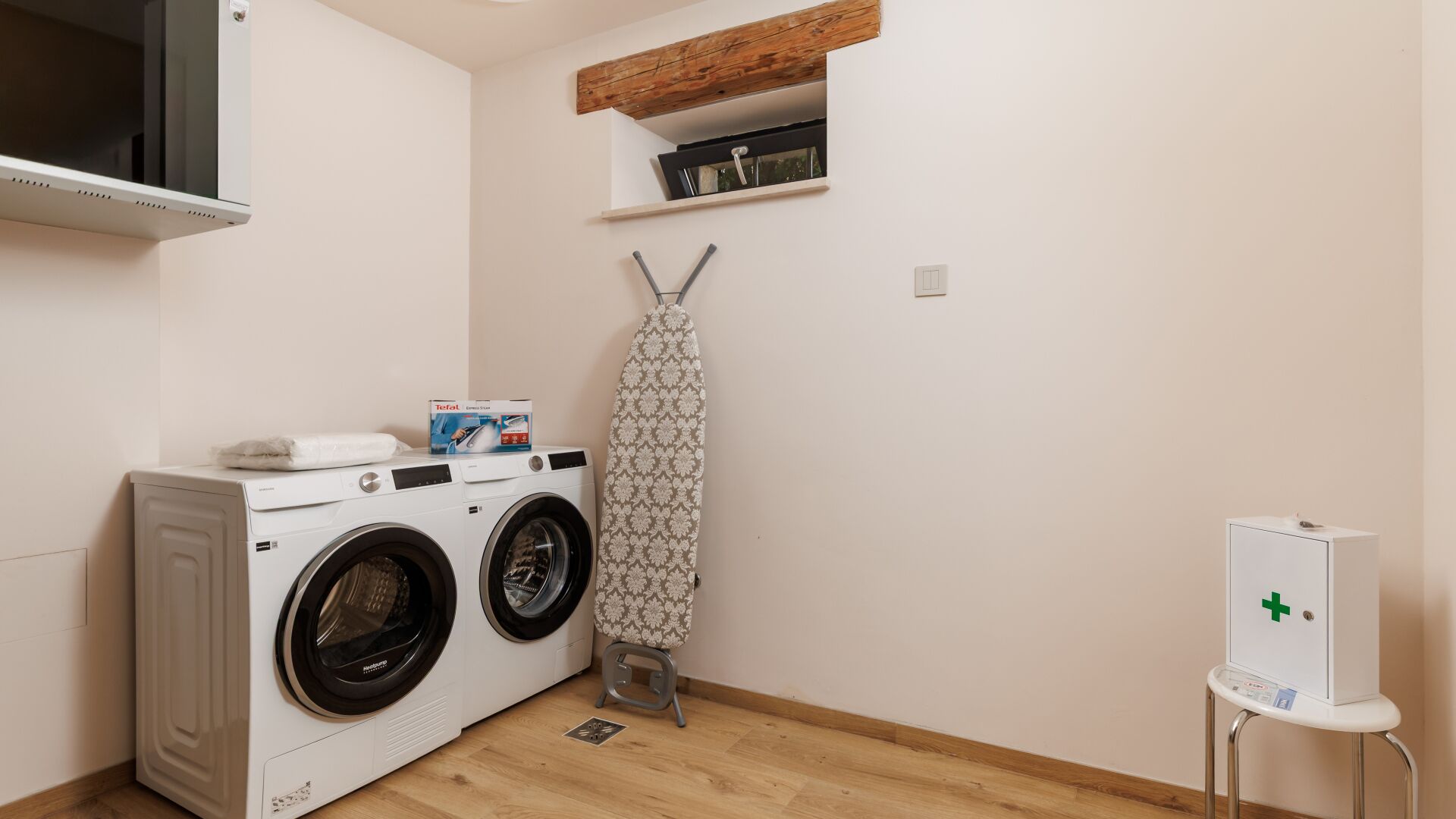 A laundry room with a washing machine, dryer, ironing board, and a small medical cabinet.