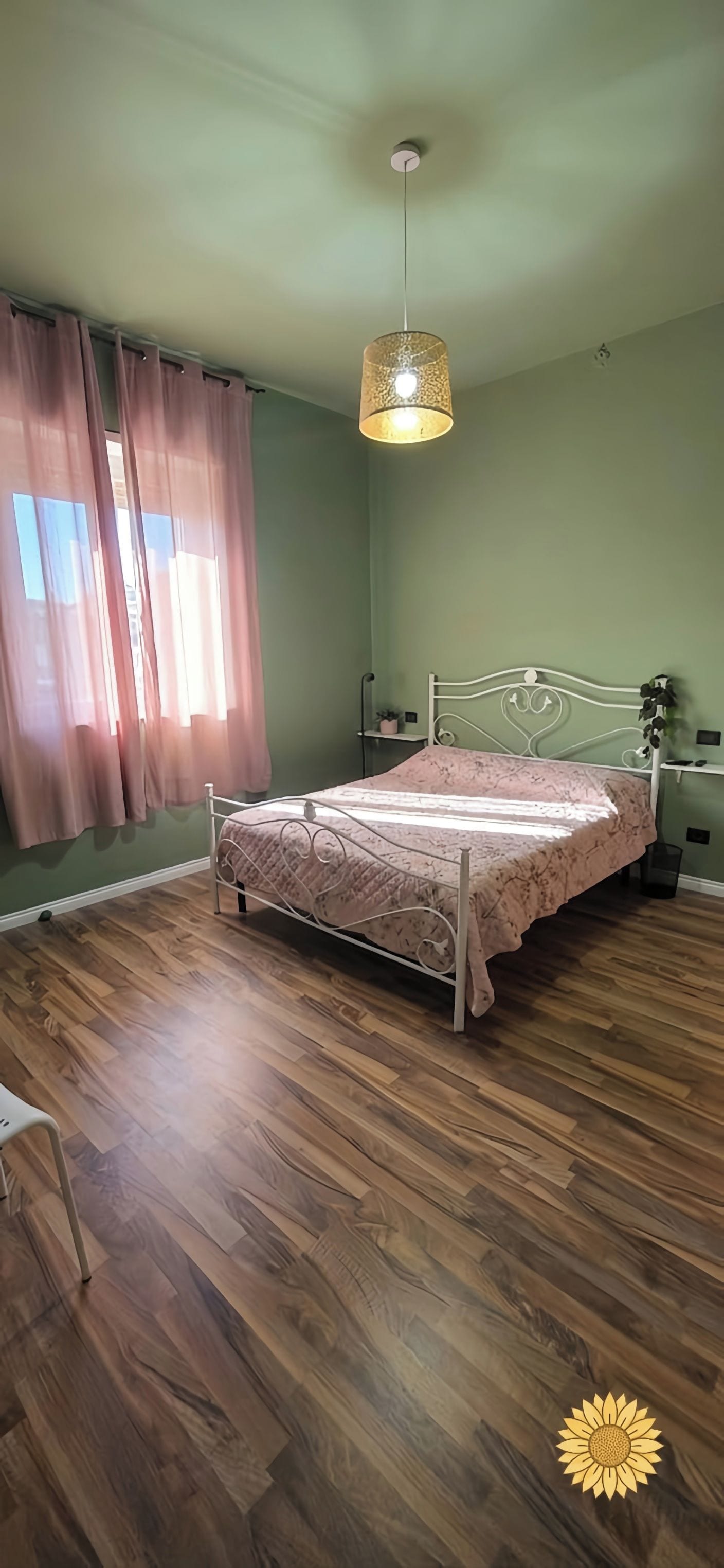 Ein Zimmer mit einem Bett, einer Hängeleuchte, einem fenster mit Vorhängen und einem Holzboden.