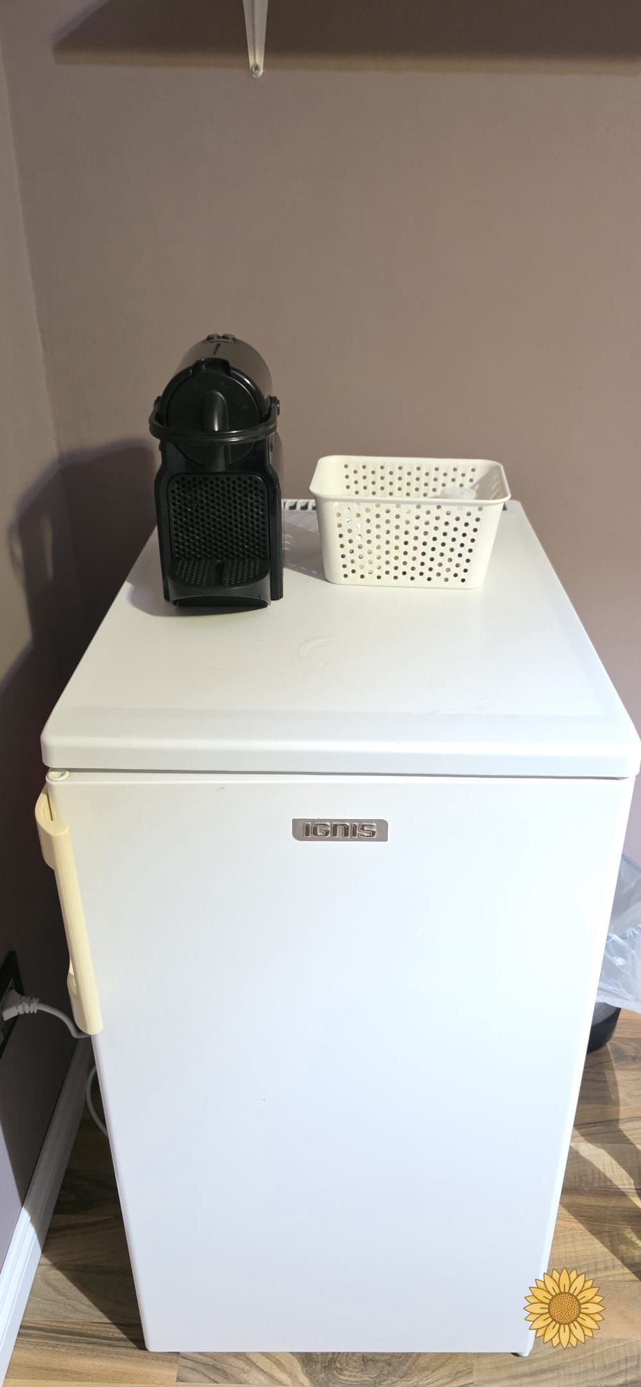A white refrigerator with a logo on the front, a black appliance on top, and a white basket beside it.