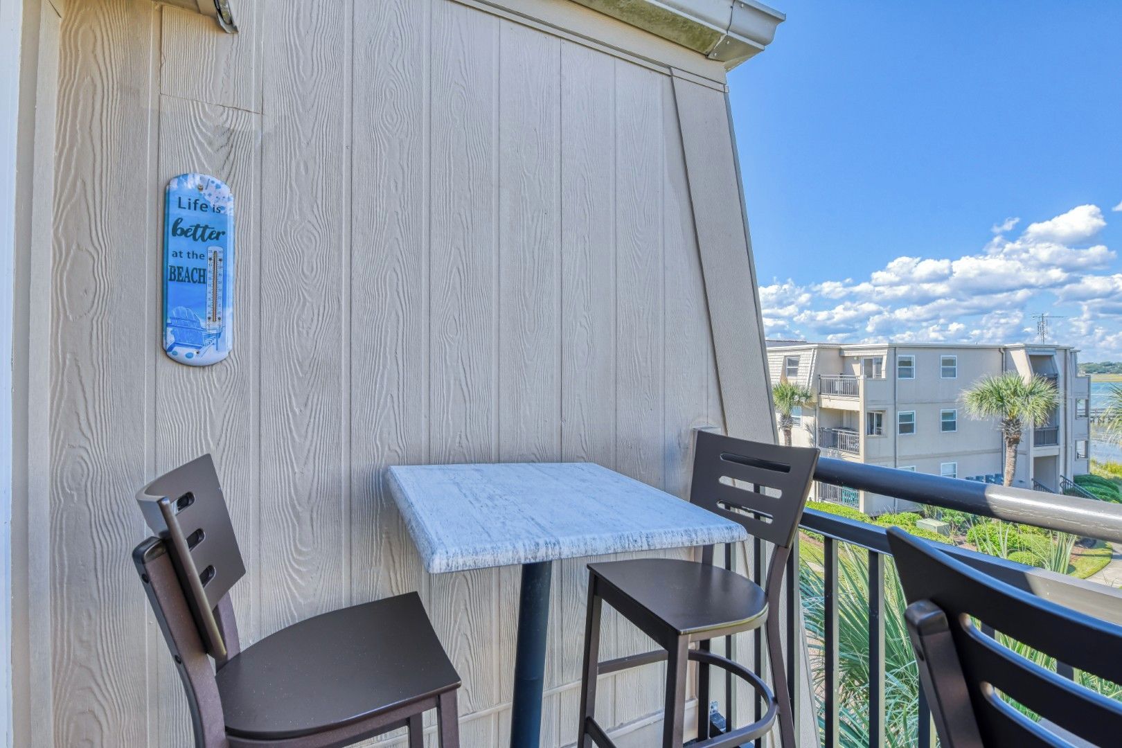 A balcony with a table and two chairs, a sign on the wall, and a view of buildings and greenery.