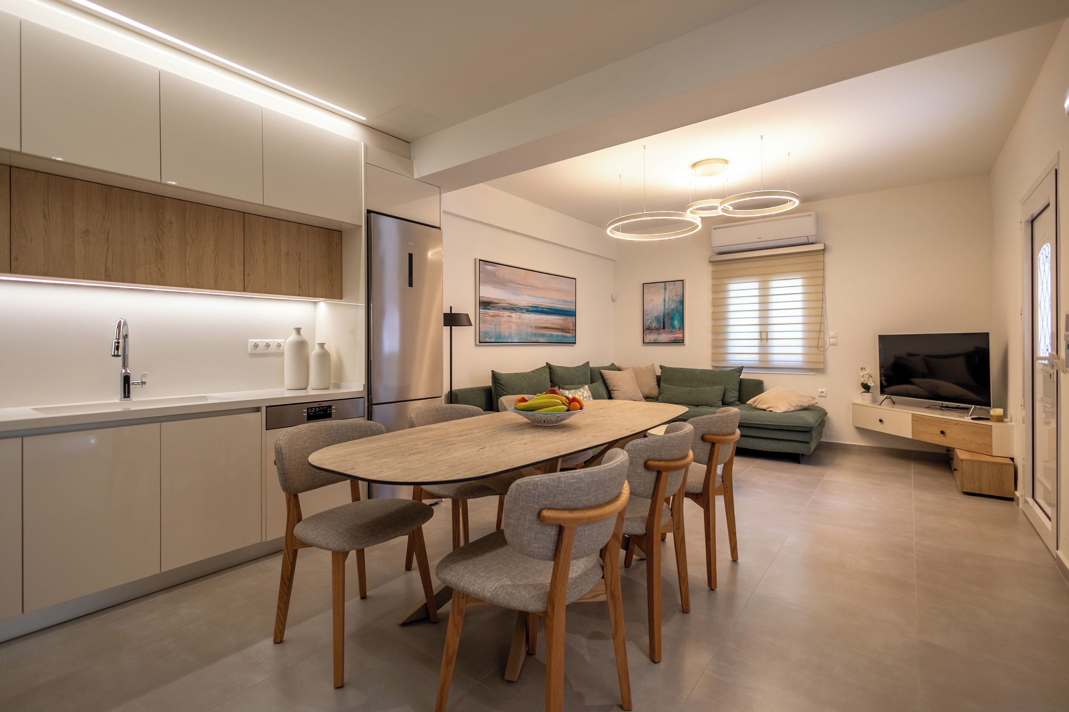 A modern kitchen with built-in appliances, a dining area with a wooden table and chairs, and a living space with a sofa and a flat-screen TV.