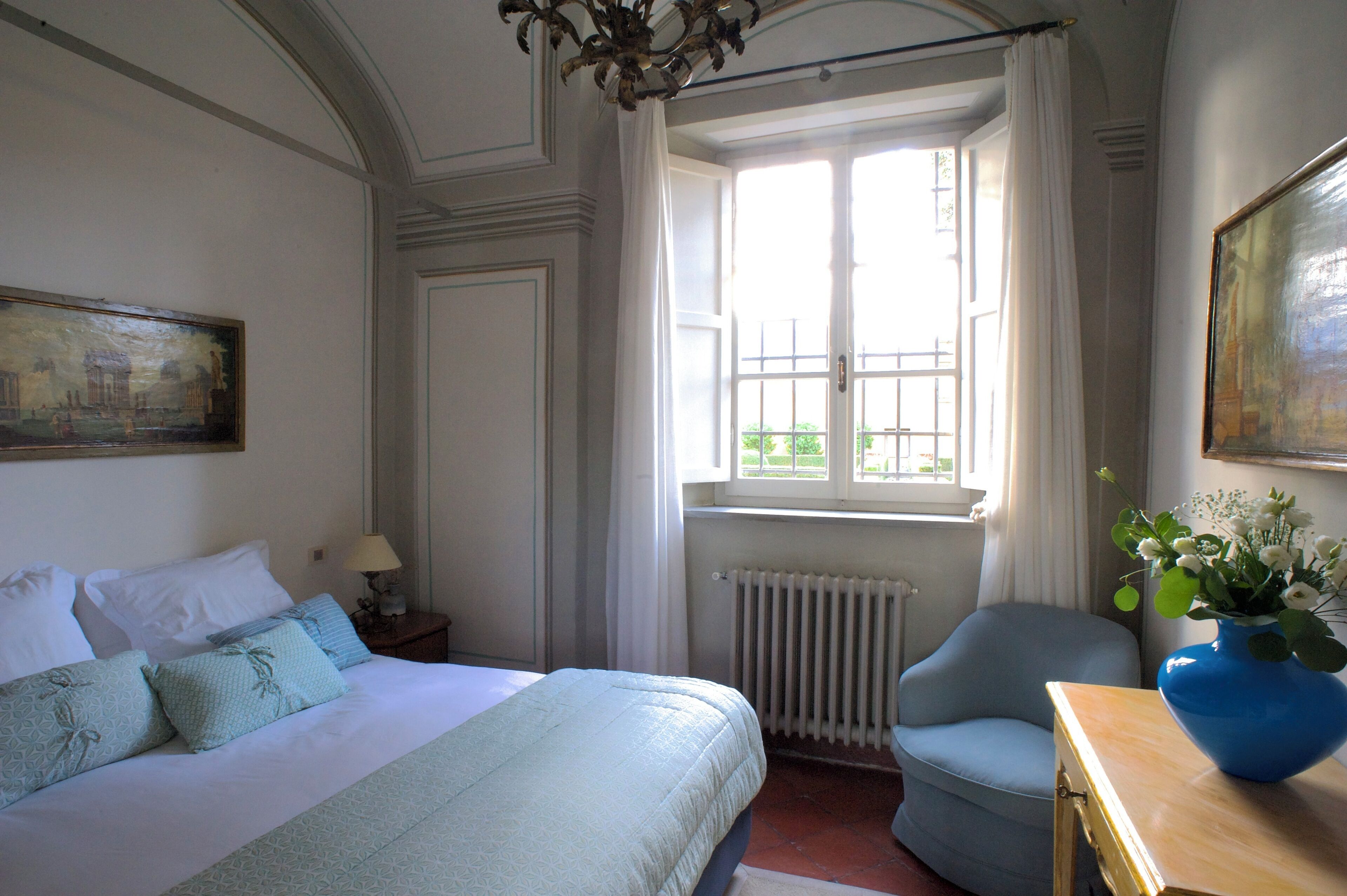 Habitación de hotel con cama, una silla, radiador, ventana con cortinas y un cuadro en la pared.