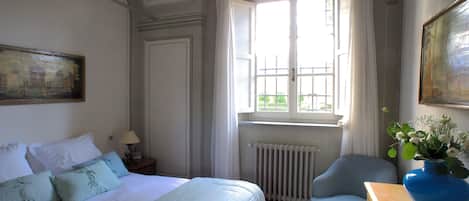 Une chambre d’hôtel avec un lit, une chaise, un radiateur, une fenêtre avec des rideaux et un tableau au mur.
