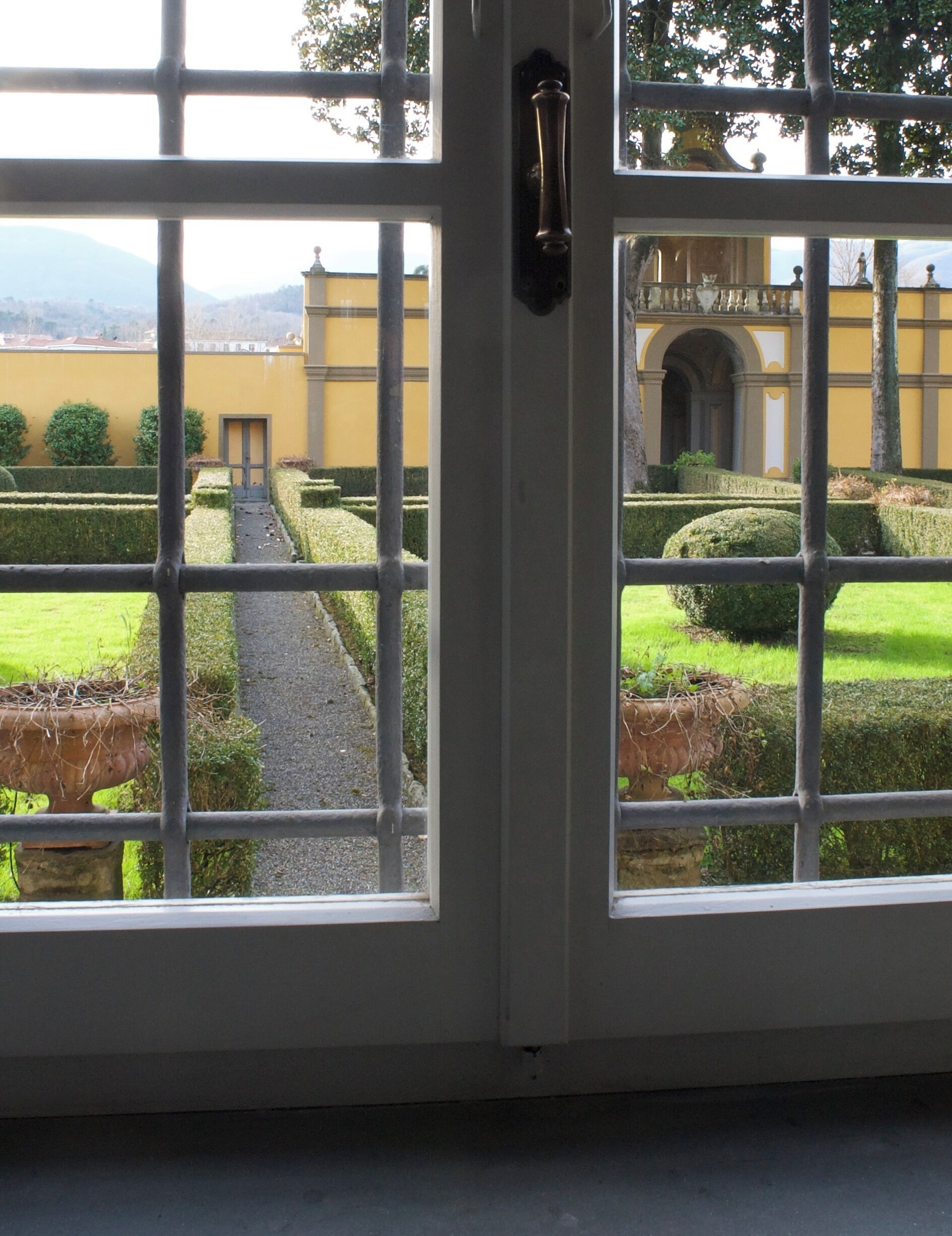 Vista a través de una ventana con reja de metal, que muestra un jardín formal con setos cuidados y un camino que conduce a un edificio de arquitectura clásica.