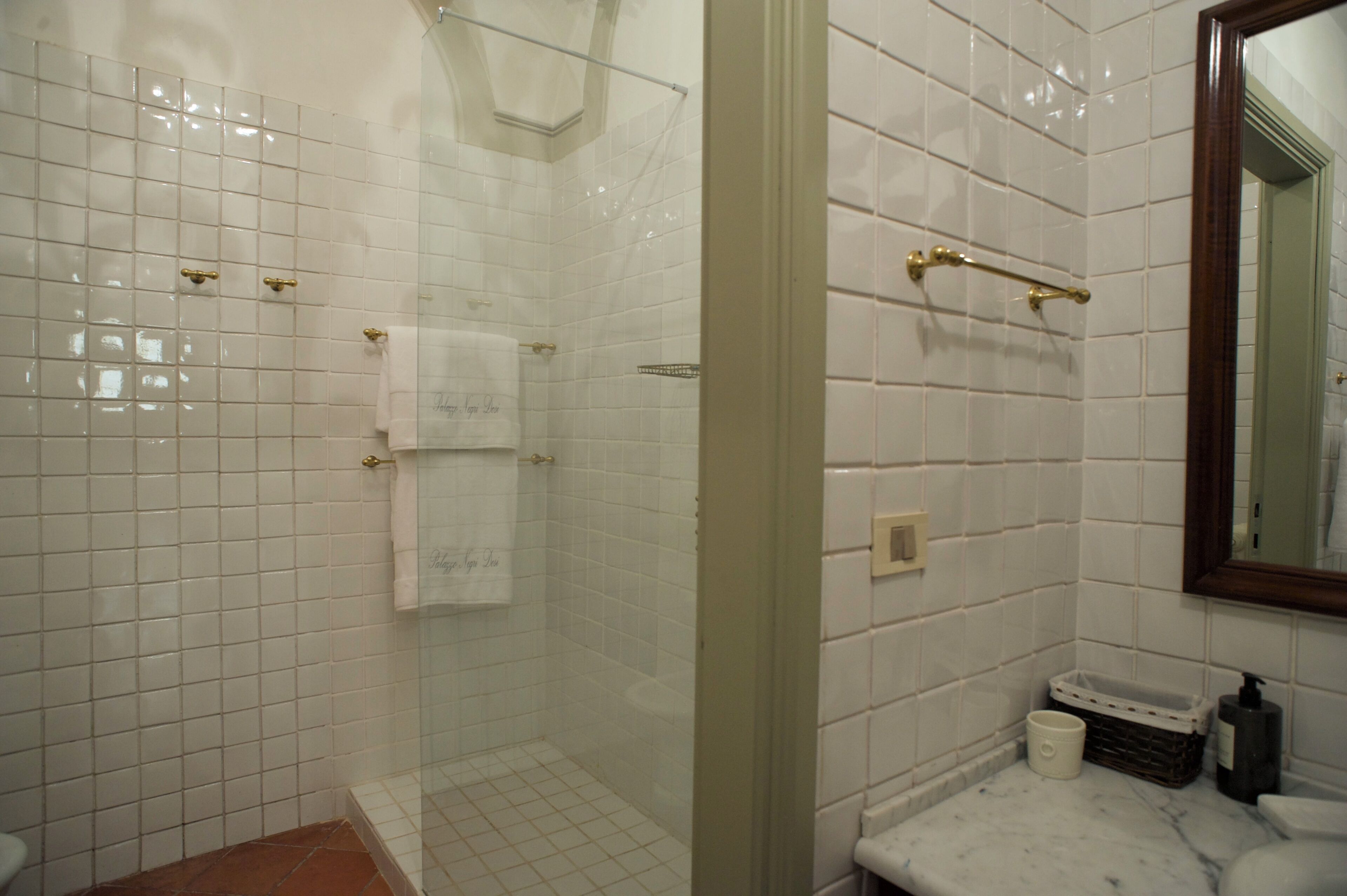 Un baño con paredes de azulejos blancos, cabina de ducha de cristal, encimera de mármol y un espejo.