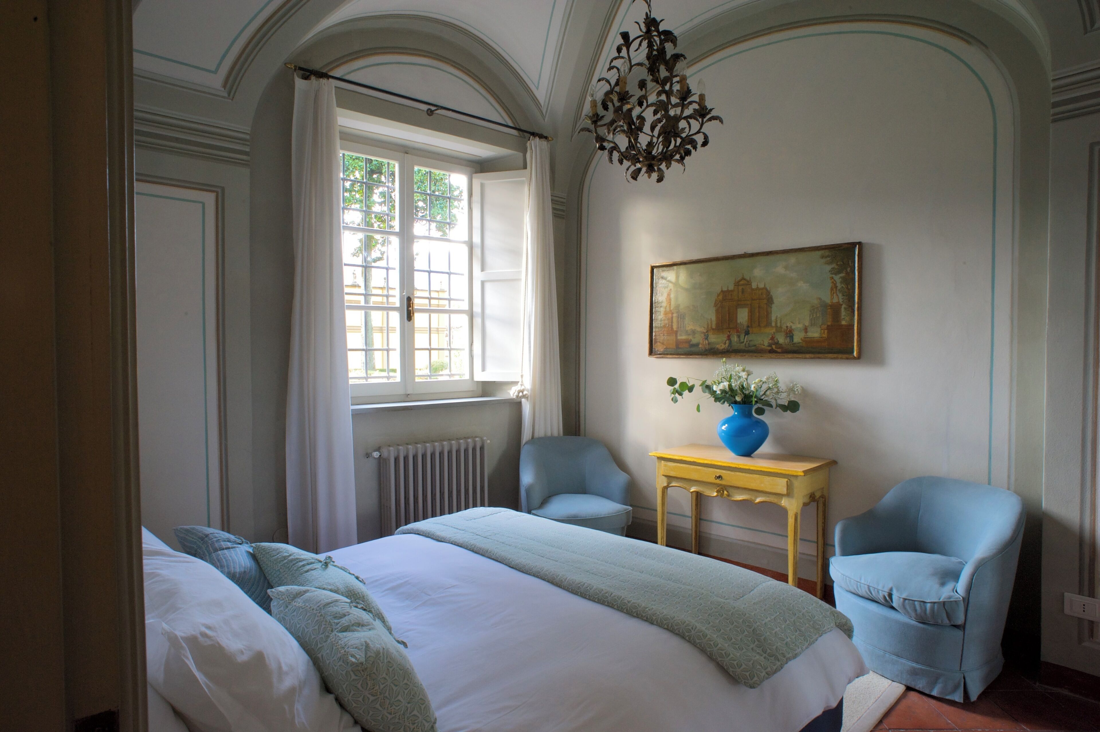 A hotel room with a large bed, two armchairs, a small table with a vase, and a painting on the wall.