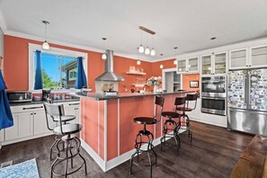 A modern kitchen with a central island, bar stools, stainless steel appliances, and a view to the outside. - 6BR Farmhouse Sleeps 14 | Fire Pit + Games + Trail (Traverse City)