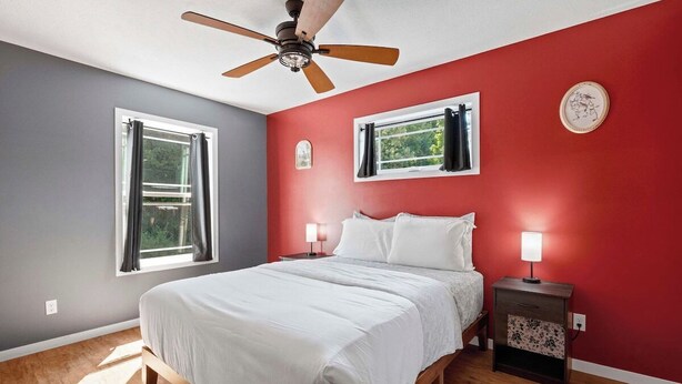 Quarto com cama, duas mesas de cabeceira, um ventilador de teto e duas janelas com cortinas.