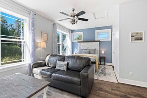 A room with a leather sofa, a bed with a headboard, a ceiling fan, and a window with curtains. - 6BR Farmhouse Sleeps 14 | Fire Pit + Games + Trail (Traverse City)
