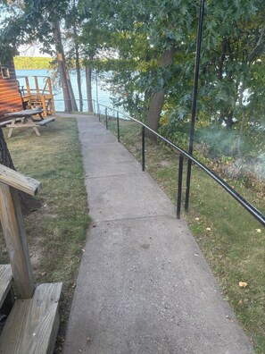 Un chemin en béton mène à une terrasse en bois dotée d’une rambarde en métal, le tout étant entouré d’arbres et bordé par une étendue d’eau.