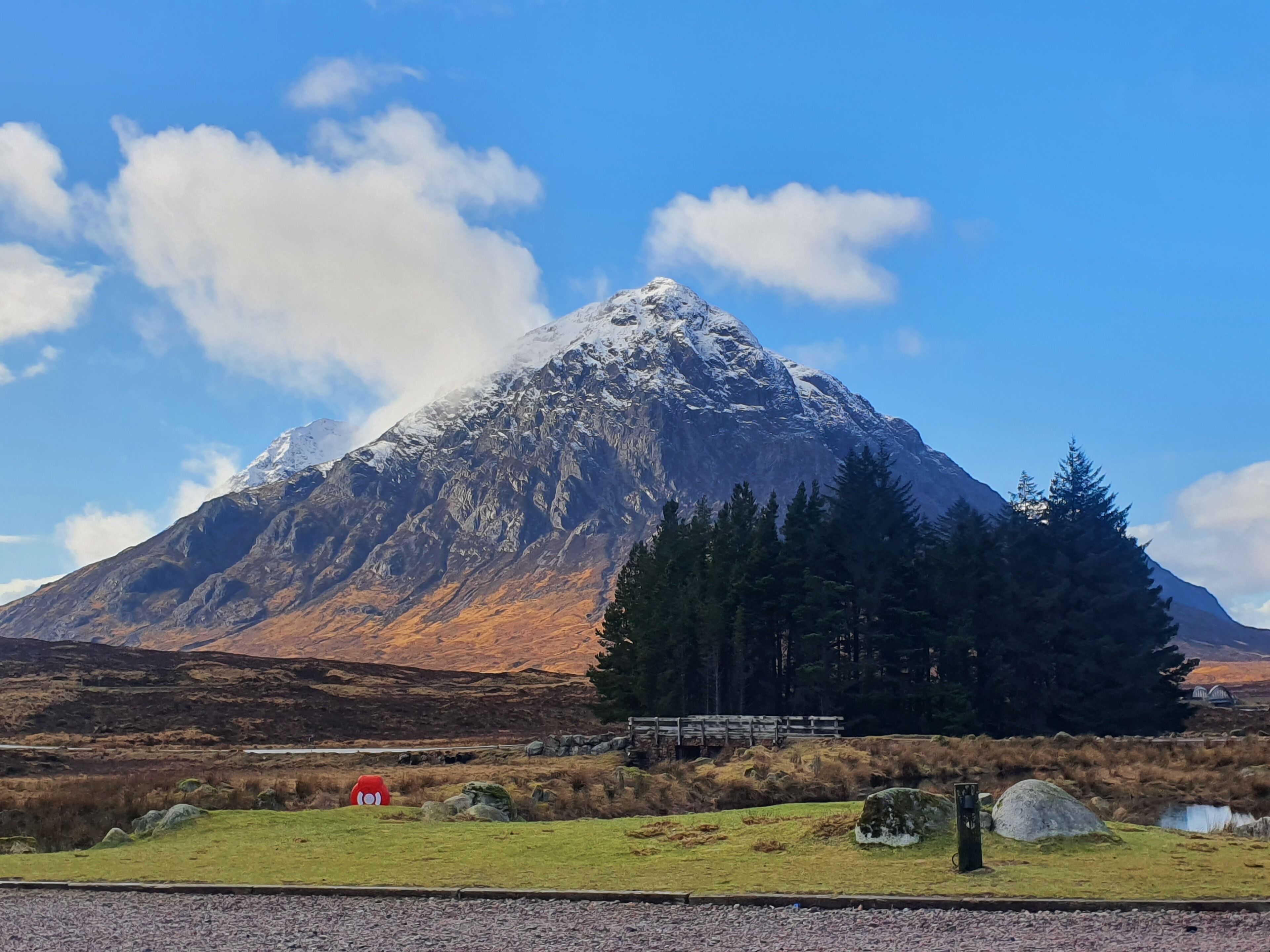 A mountain with a snow-capped peak, a grassy foreground with a red object, and a wooden bridge.